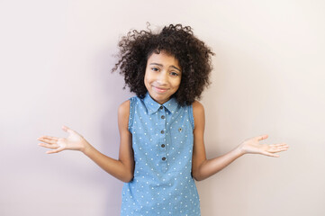 Portrait of cute biracial preteen girl shrugs shoulders, a schoolgirl wearing polka dot shirt looks at camera spreads her hands as a sign of misunderstanding, incomprehension, isolated on pastel pink