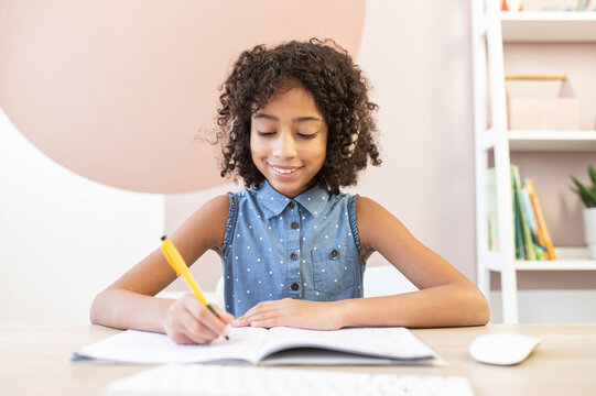 A portrait of a pretty curly African schoolgirl wearing a headphones, a pretty biracial girl fill exercises book in with a pen, doing home work. Concept of studying on the distance