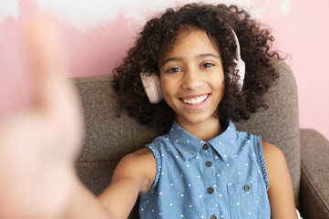 A cheerful curly African-American preteen girl wearing a headphones is taking a selfie. A close-up portrait of pretty biracial school-age girl blogger
