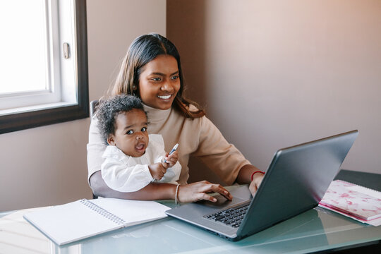 Mixed race Indian mother with African black baby working online from home on Internet. Workplace of freelancer woman with kid. Stay home single mom working distant job. A new normal.