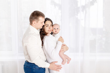 family, the concept of a happy young family with a small child, father, mother and daughter hugging at home by the window