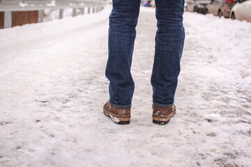 Male legs in blue jeans and boots on snowy sidewalk. City people walk in winter. Pedestrian back view, copy space