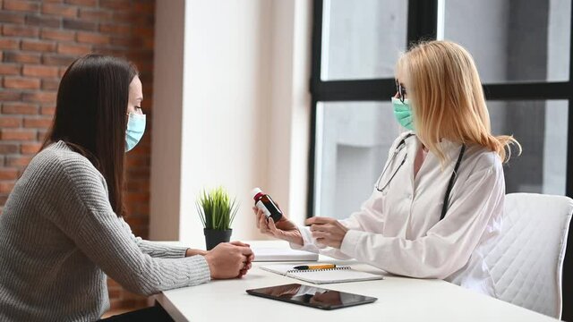 A female patient and a doctor wearing masks in the hospital office. A female MD with a bottle of remedy in hand prescribes treatment, recommends medicine. Appointment during virus outspread