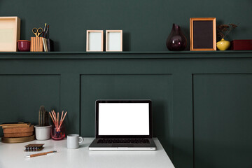Computer with white screen on a desk with drawing supplies, coffee mug, frames on cactus green paneled wall.