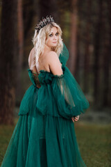 young beautiful blonde hair woman queen. Princess walks. autumn green forest mystic. Vintage medieval shiny crown. Long evening green dress. magic fantasy. back view