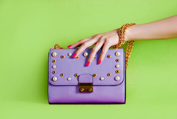 Purple purse handbag on green background. Woman hand with red manicure touching it 
