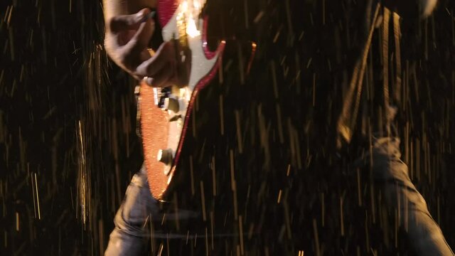 Rock musician plays a red white electric guitar in a dark studio against the background of falling rain drops. Close up of male hands play on strings with guitar pick. Slow motion.
