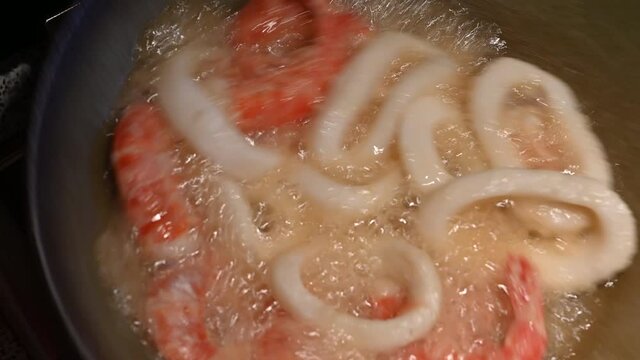 Home life scene: close-up footage of shrimps and floured squid rings being fried in hot oil. By moving the pan, they are made to rotate inside to facilitate cooking.