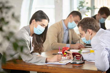 Side view colleagues working together in a office using modern devices during creative meeting. Stationery, laptop, documents. Concept of business, office, finance, open space. Wearing face mask.