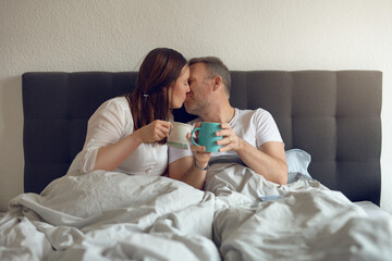 Happy middle-aged couple kissing in bed in the morning holding a mug of coffee