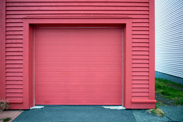 A large bright red garage door surrounded with wood clapboard siding. The building has asphalt paved entrance to the door with grass on the side of the building. There's a white building on the side. 