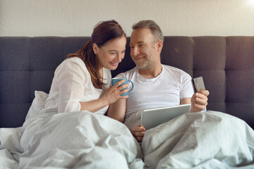 Happy middle-aged couple sitting in bed in the morning drinking coffee and doing online shopping with a digital tablet