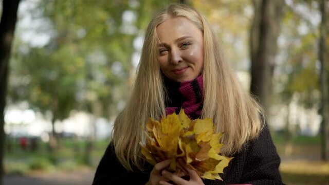 Portrait of happy charming Caucasian young woman posing with leaves bouquet in autumn park. Smiling beautiful slim lady looking at camera standing outdoors in sunrays. Joy and beauty concept.