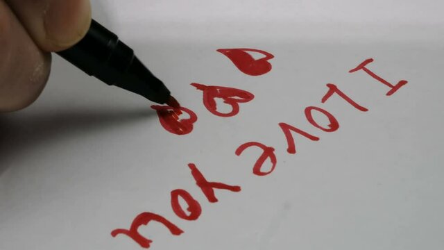 A young guy draws a valentine card for his girlfriend. Close-up of a young man's hand he writes on paper the words I love you with a red pen.