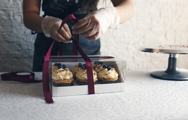 A girl cook in a gray apron packs cupcakes with cream in a gift box to send the order to the...