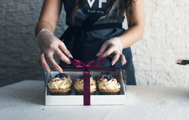 A girl cook in a gray apron packs cupcakes with cream in a gift box to send the order to the...