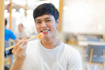 close up young cute asian japanese man smiling while using chopsticks to hold freshness sliced salmon (sushi) at restaurant for asian eating culture tradition concept