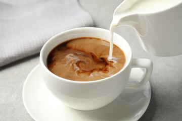 Pouring milk into cup of hot coffee on grey table, closeup