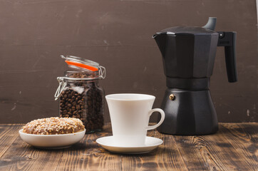 Cup of cofee with cofee beans and black maker. Selective focus. Cup of black coffee in a cup, donut with nuts and grains in a glass jar with a coffee maker on a brown background.
