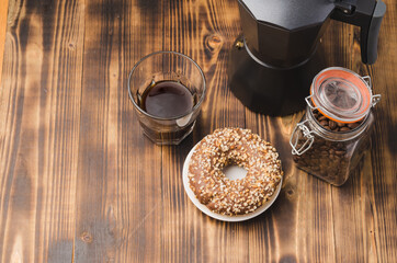 Cup of cofee with cofee beans and black maker. Top view. Cup of black coffee in a cup, donut with nuts and grains in a glass jar with a coffee maker on a wooden background.
