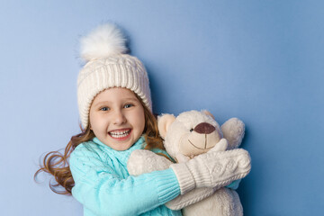 happy little 5-year-old girl with blonde hair in a knitted sweater cap and mittens, smiles and hugs teddy bear lying on blue background in the studio. toy is her friend. Winter time. Holiday weekend