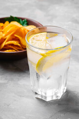 Fresh ice cold water drink with lemon near to fried corrugated golden potato chips with parsley leaf in wooden bowl on concrete background, angle view