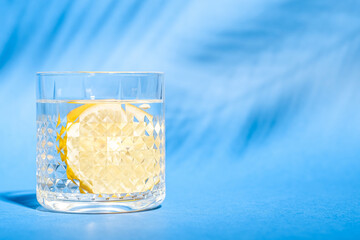 Glass with water and lemon on blue background with tropical palm leaf shadow