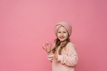 little girl in a fluffy sweater and a headband, smiling with a doughnut in her hand. Sweet appetizer, delicious pastries. a child shows a delicious dessert on a pink background in studio
