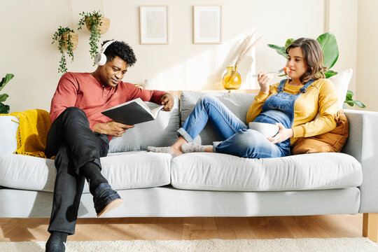 Pregnant wife eating on the couch with her husband who is listening to music and reading a book. Interracial couple concept