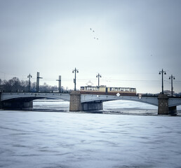 A tram rides over a concrete bridge in winter, a frozen river with white snow and ice. Over the bridge, flying ducks.Winter in St. Petersburg