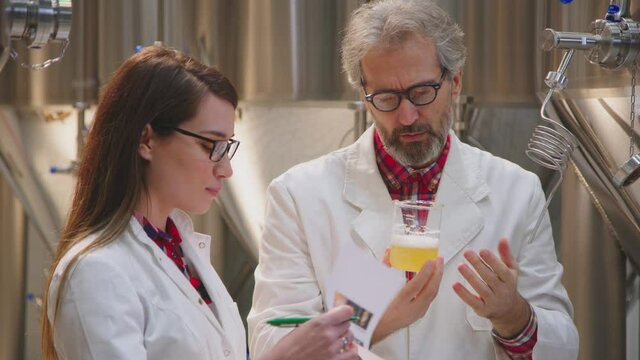 Two scientists wearing glasses, talking about beer sample and taking notes. Food technologists testing beer quality. Science, brewery concept.