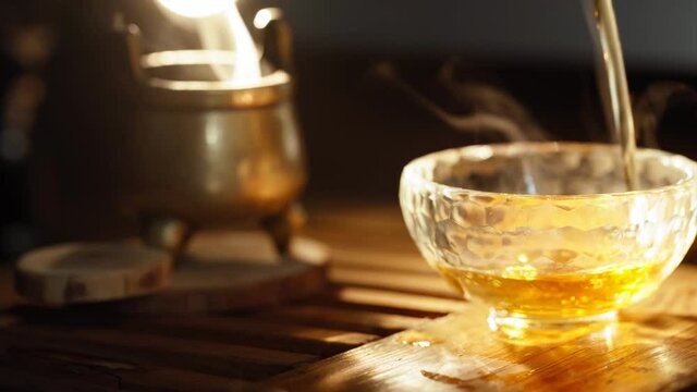 Herbal Chinese Tea Poured into Beautiful Transparent Glass against background of the smoke coming from scented sticks, backlit by warm light. Tea ceremony. Oriental cup close up. Unrecognizable person