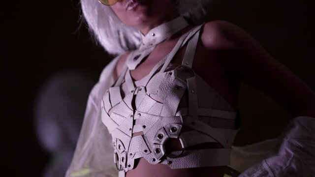 Portrait of a blonde woman in stylish glasses in an unusual white sexy strappy suit posing in a studio against a dark background.