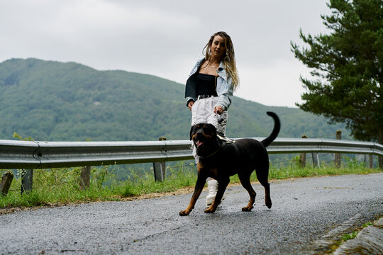 Young woman with blonde braided hair wearing a denim jacket and white jean walking her dog on a rainy day