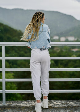 back side of a young woman with blonde braided hair wearing a denim jacket and white jean resting on a dam