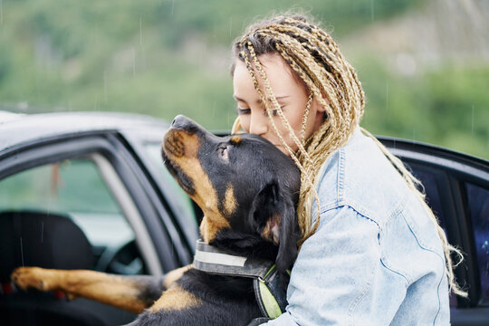 Young woman with blonde braided hair wearing a denim jacket and white jean kissing her dog on a rainy day