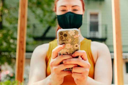 Young woman sitting outdoors texting on phone, wearing face mask.