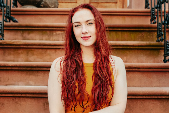 Portrait of a young woman with red hair sitting outdoors on a stoop.