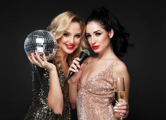 Two beautiful women, blonde and brunette in evening dresses, chic hairstyles and makeup, sing with...