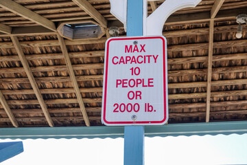 White sign with red letters on a pole with the inside of the wooden ceiling as a background. The sign says Max Capacity 10 People or 2000 lbs