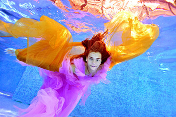 A beautiful young girl is swimming underwater in a pool in a purple dress and with a yellow cloth in her hands. Her arms are spread out like wings. Underwater fashion photography.