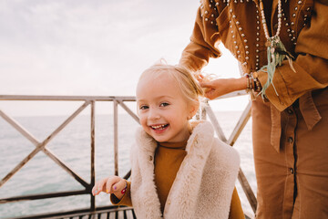 Baby girl smiling next to the sea promenade. Baby girl with blonde hair, in a warm fur vest. Spring time, sun is shining on her face. Walk along the seafront with family. Travel abroad. 