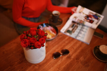 Young white brunette girl in red is having breakfast in a cafe. She was presented with flowers on a holiday. She drinks tea or coffee, eats a bowl of fruits and cereals, and reads a fashion magazine.