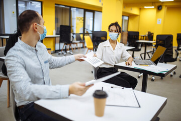 Young colleagues pass documents to each other working at an office during coronavuris pandemic outbreak. Professional workers wearing medical masks checking news and trends in business.