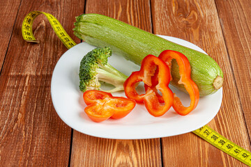 a white plate with a zucchini, broccoli and some pieces of Bulgarian pepper standing on a yellow centimeter on brown wooden background