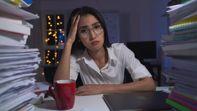 Serious asian business woman is sitting at desk covered with stack of paperwork and looking camera