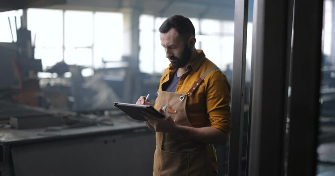 Factory worker checking the quality of metal products, writing on a digital tablet. Examination of metal doors at the factory. Use of digital technologies in the production