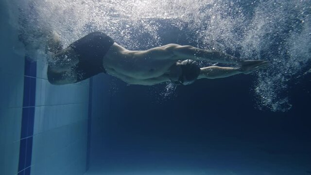 Cinematic shot of young professional male swimmer with cap and goggles trains with effort and dedication to win the race by practicing in swimming pool. Concept of sport, competition, victory, fitness