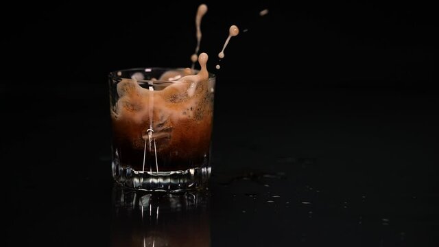 Multiple ice cubes drop in a glass of cola, splashing and foaming drink in slow motion
