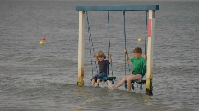 Two children sit on wooden swing above sea, children play, splash sea water with feet